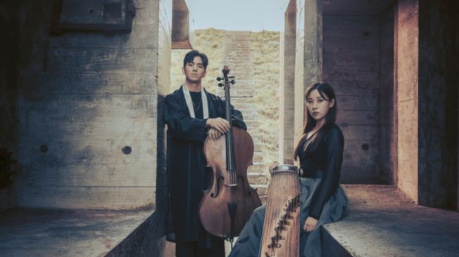 Photo of o woman with a wooden musical instrument and a man with a cello. They are standing in a narrow corridor between concrete walls.