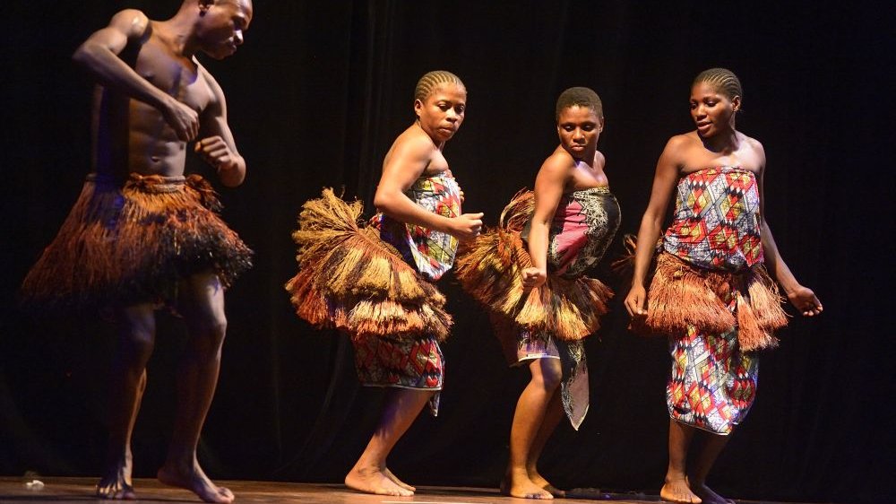 Photo of four Africans dancing on a stage - three women and a man in colourful, ethnic clothes.