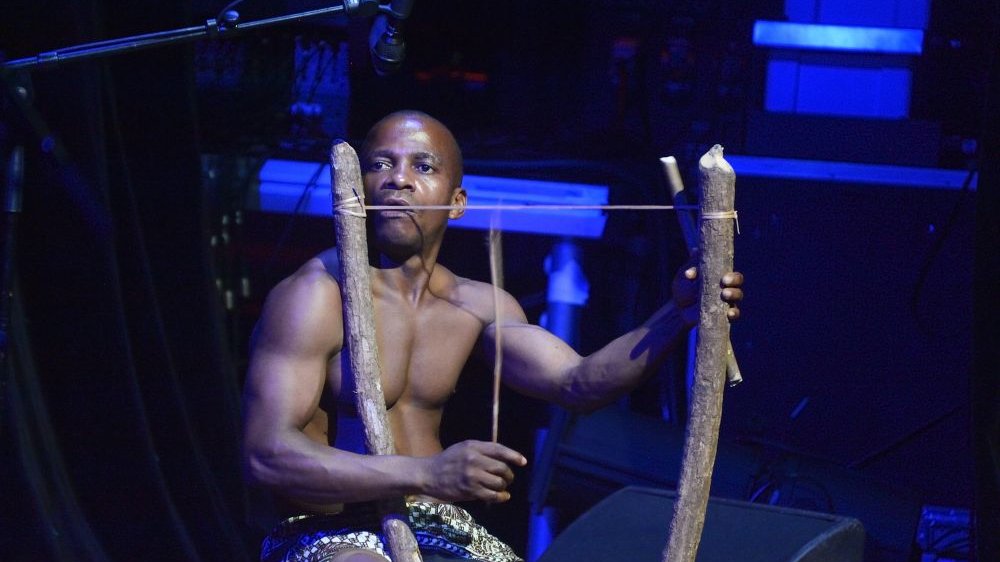 Photo of the African man sitting by a simple wooden instrument. Dark background.