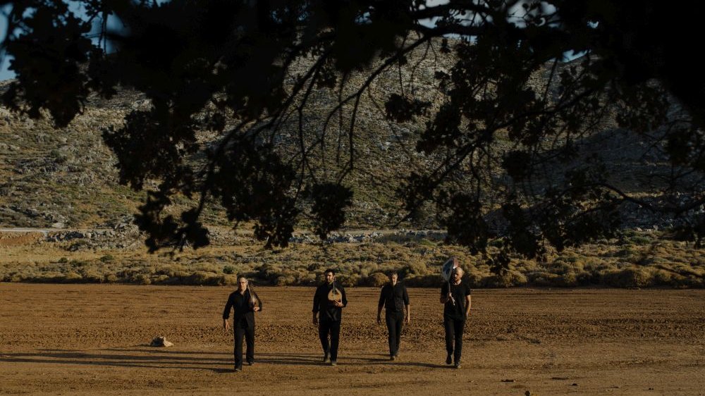 Photo of four men, three of them are holding musical instruments. They are going through the open space (a field). Tree branches in a foreground and low bushes as a background.