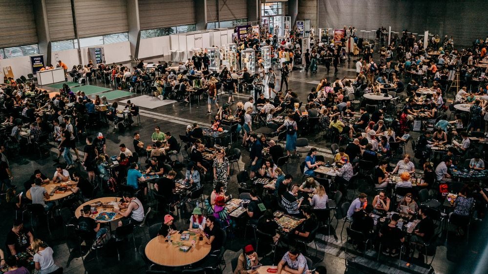 Interior of an exhibition hall: many people sitting at the tables and playing games.