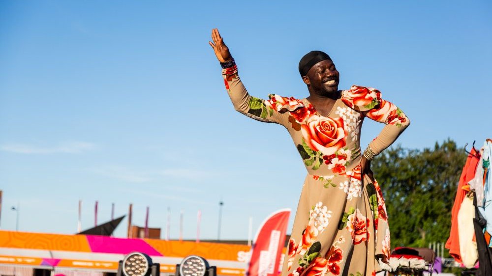 Photo of a smiling Afro-American dressed in a floral dress, with one of his hands raised, and the other on his hip. Blue sky and a tree as a background. - grafika artykułu