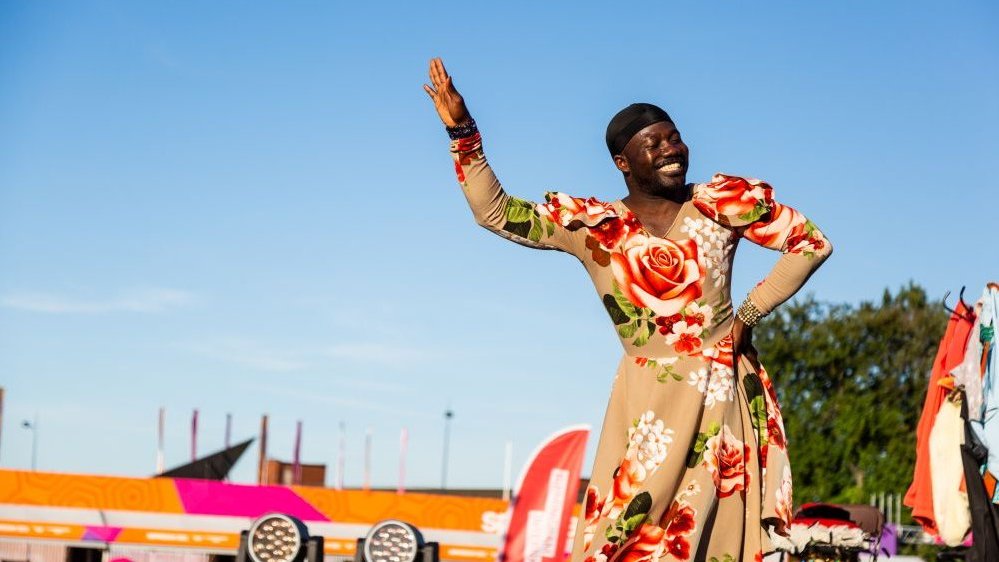 Photo of a smiling Afro-American dressed in a floral dress, with one of his hands raised, and the other on his hip. Blue sky and a tree as a background.