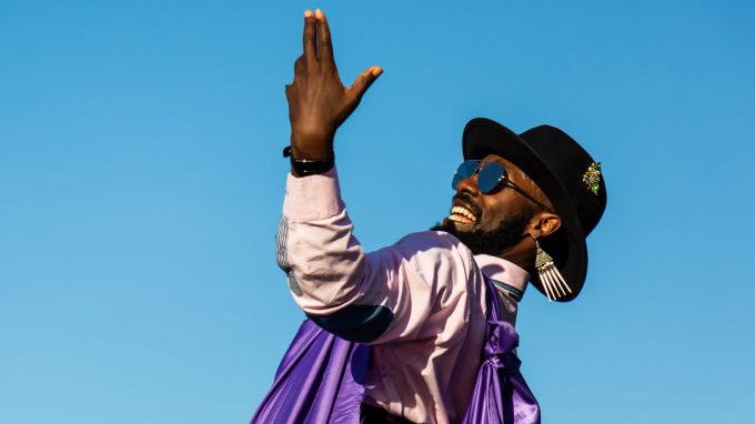 Photo of a smiling Afro-American, in sun-glasses and a black hat, The man dressed in white shirt and a violet shawl is standing sideways with his one hand raised. Blue sky as a background.