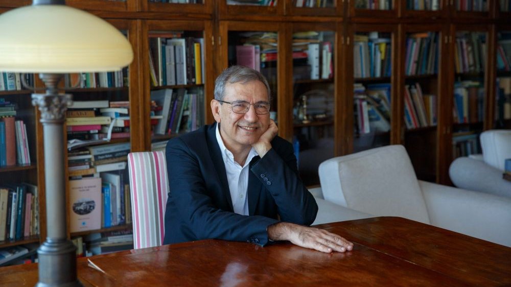 Photo of smiling Orhan Pamuk, who is sitting at the table. The bookcase as a background.