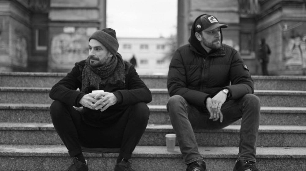 Black and white photo of two men, who are sitting on the stairs outside. One of them is holding a disposable cup.