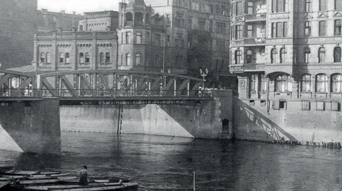 Black and white photo of the river, a few boats, the bridge and buildings along the river.
