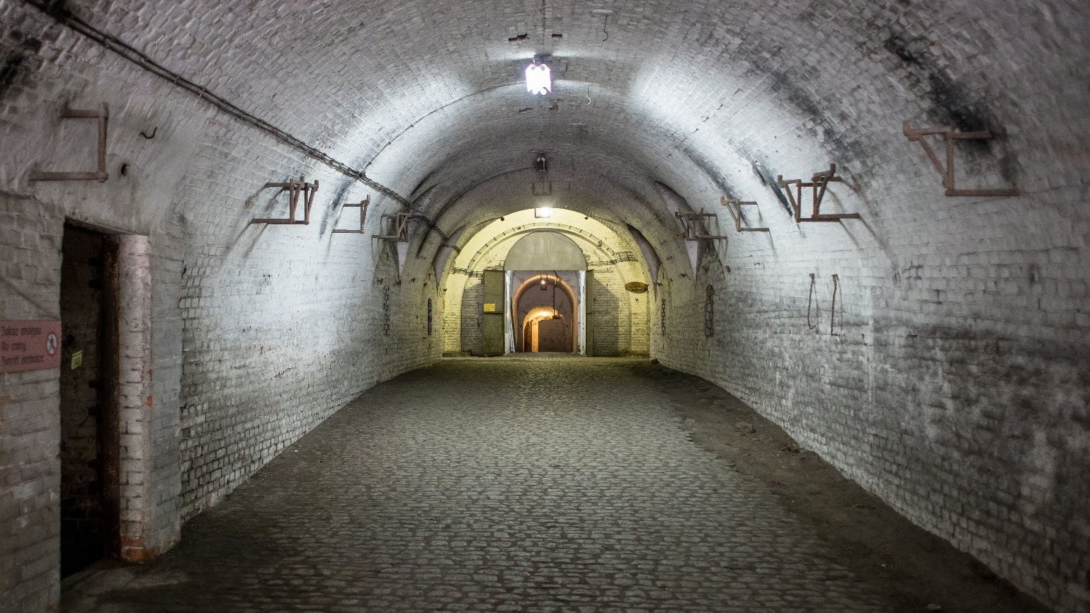An underground, wide corridor, illuminated by artificial light. On the left an entrance.