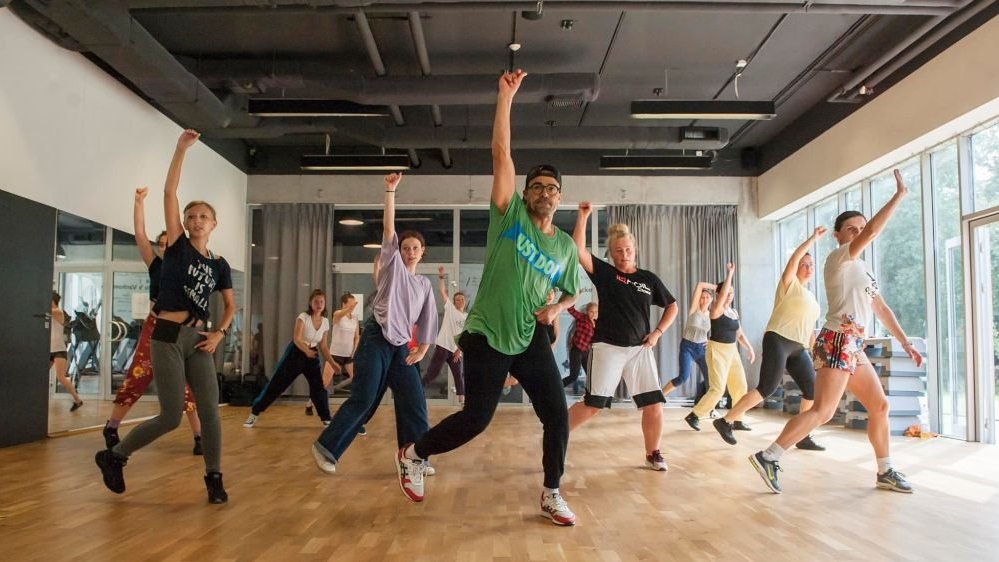 A group of dancing people in a big room, all of them in similar poses, with one hand stretched up.