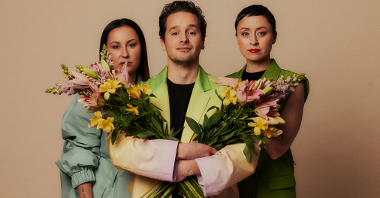 A young man standing between two young women and holding two bunches of flowers.