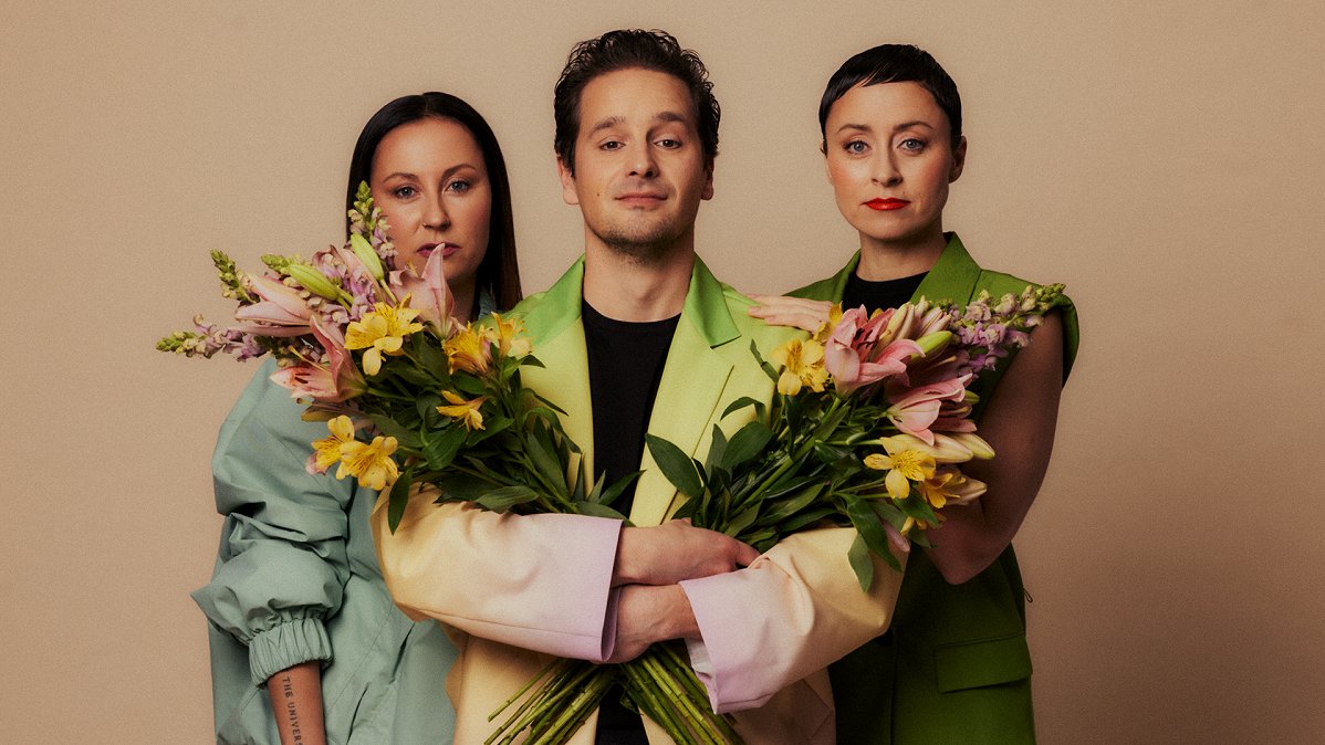 A young man standing between two young women and holding two bunches of flowers.