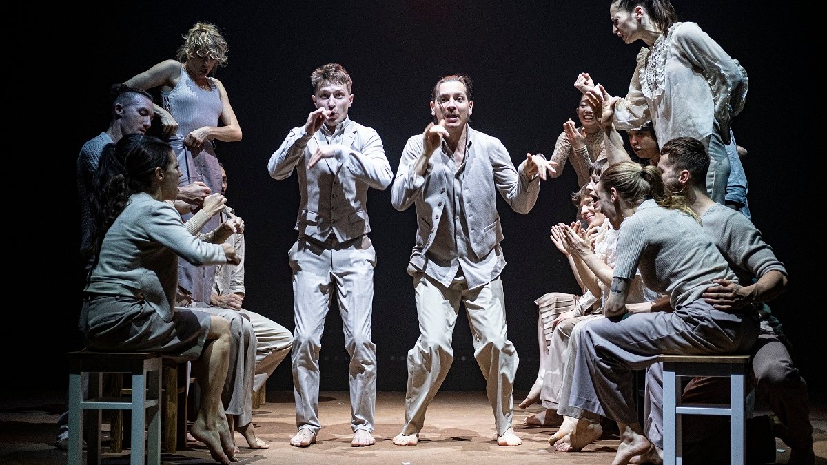 Photo of a group of dancers, some of them standing and some of them sitting on stools.