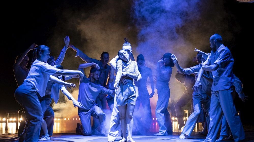 Dancers in various dance poses: a man hugging a woman in the middle and other dancers around them. The dancers are illuminated with blue light.