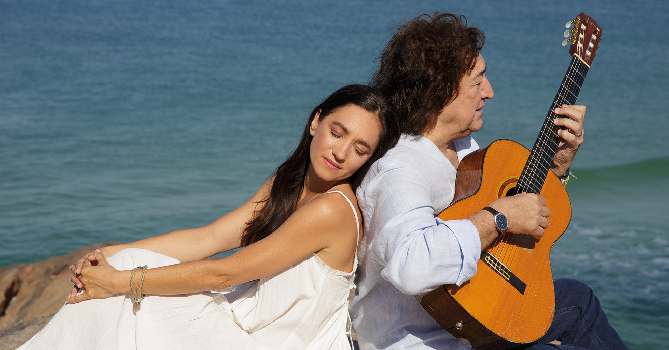 Photo of a woman with closed eyes and a man with a guitar sitting back to back; sea as a background. - grafika artykułu