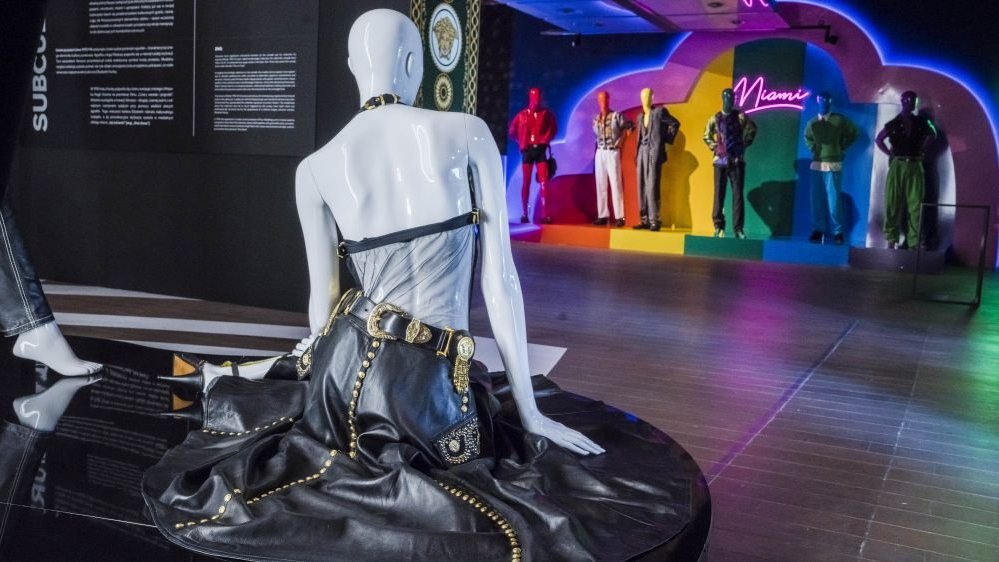 A white mannequin in a black leather dress sits backwards. In the background, mannequins in men's clothes against a colorful wall with a neon sign.