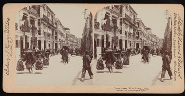 Stereophotograph of a street in sepia colours: in the foreground two people carrying goods on their shoulders. Lane of buildings on the left.