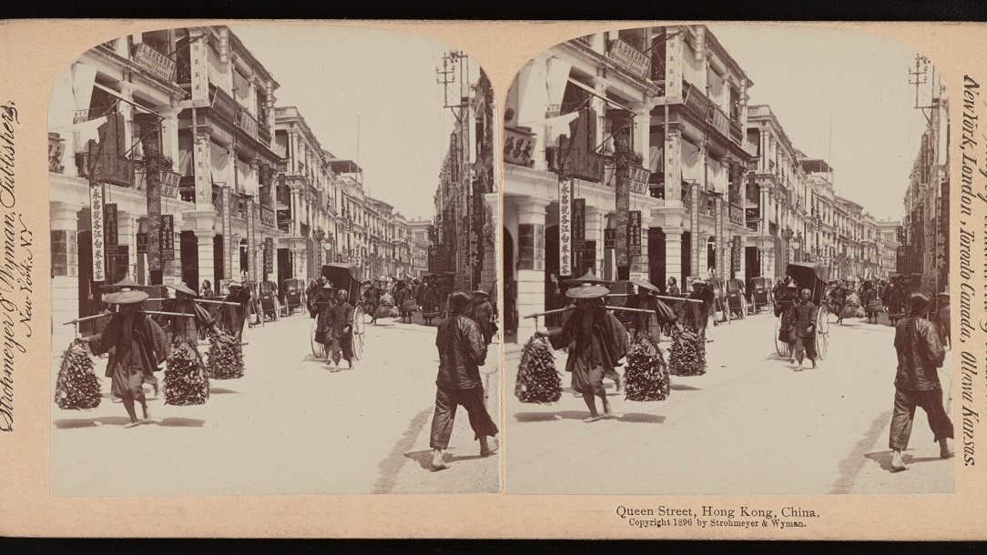 Stereophotograph of a street in sepia colours: in the foreground two people carrying goods on their shoulders. Lane of buildings on the left.