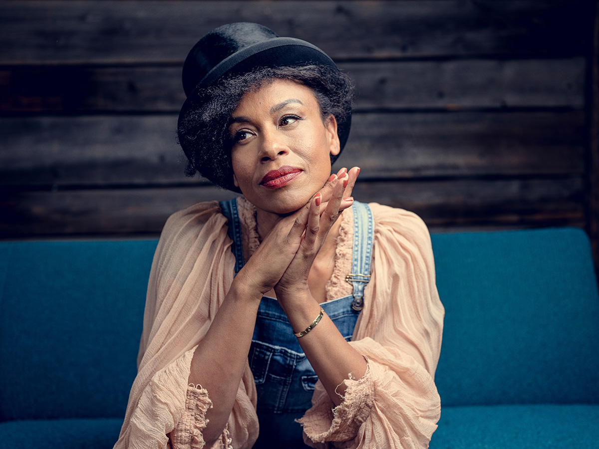 A young woman with dark hair, wearing a hat, sitting on a sofa, looks to the right. The woman has her hands folded under her chin. - grafika artykułu