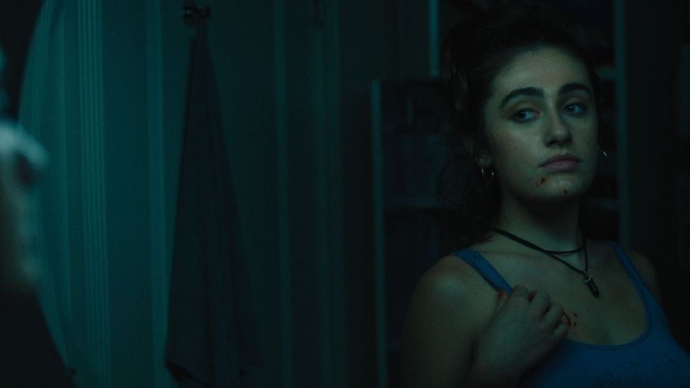 A dark room, in the corner a black-haired girl in a purple T-shirt, with a necklace. There are small traces of blood around her chin.