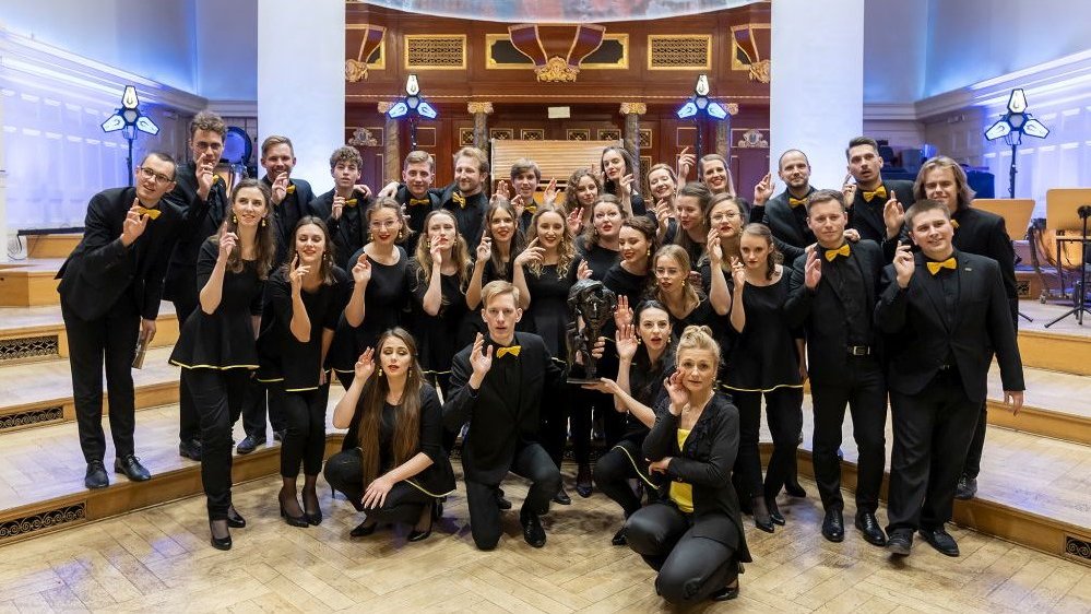 Photo of the choir members, young women and men, dressed in black clothes, standing on a stage. All of them are smiling and have their right hands raised.