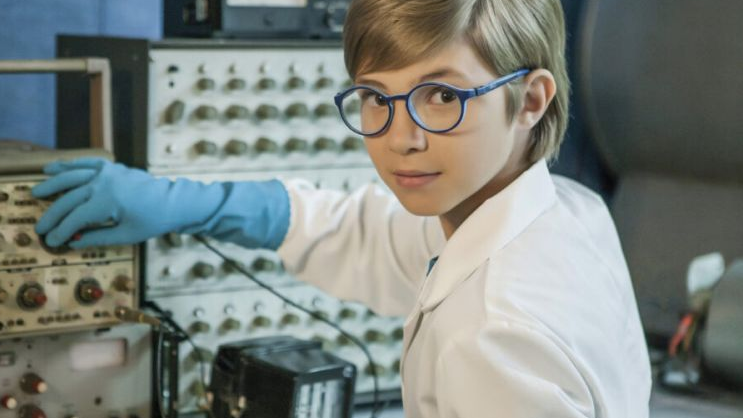 A boy in a white apron and with a rubber glove on his hand, turned towards the camera and simultaneously touches the knob of the apparatus in front of him.