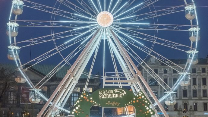 An illuminated Ferris wheel, a green gate in front of it and people walking around.