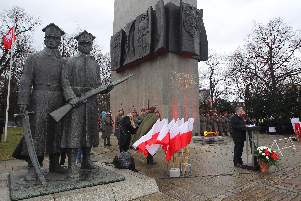 Photo of the monument to the Greater Poland Insurgents. A man speaking in front of the monument and white and red Polish flags, soldiers around the monument. - grafika artykułu