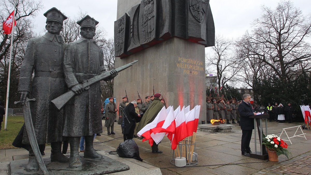 Photo of the monument to the Greater Poland Insurgents. A man speaking in front of the monument and white and red Polish flags, soldiers around the monument.