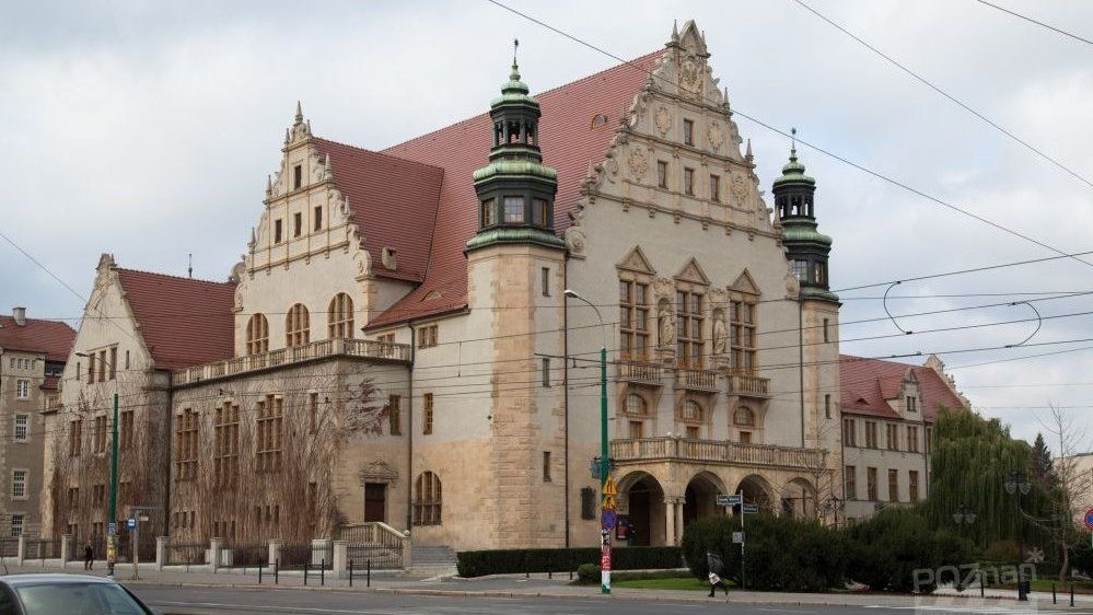 The historic building of the University Hall with two turrets and decorations on the facade. In front of the building there are bushes, a sidewalk and a street.