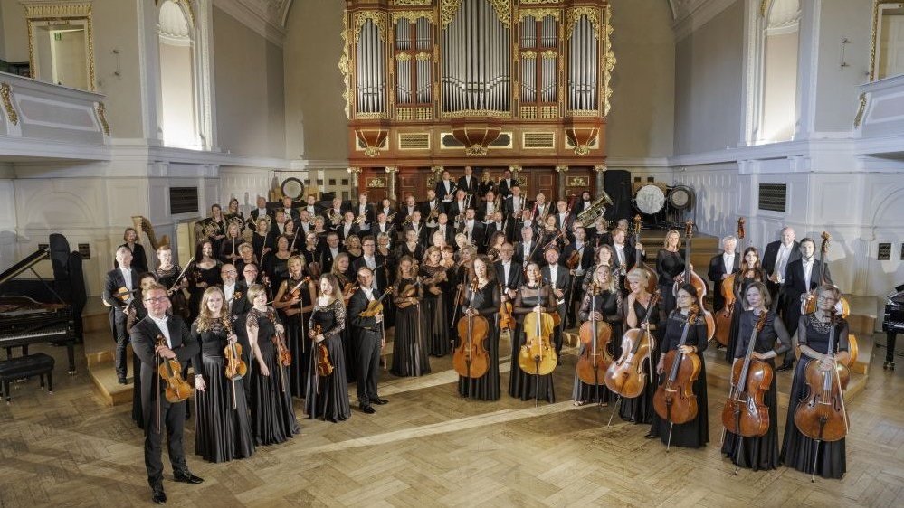 Musicians with their musical instruments, standing on stage in semicircle. Pipe organ in the background.