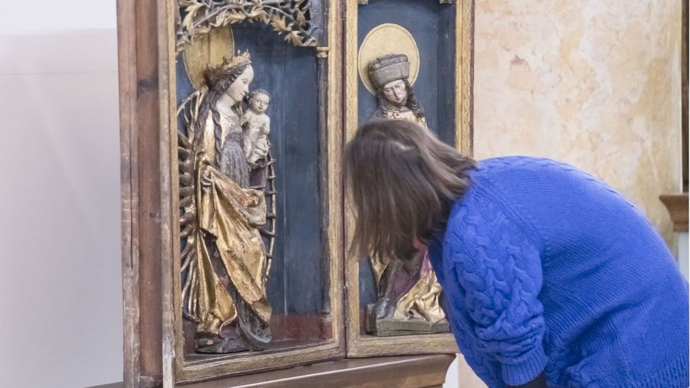 A young woman visiting the exhibition leans over the exhibit - a wooden altar closed in a display case, on which there is a figure of the Virgin Mary with Jesus. The figure is richly decorated and full of gold.