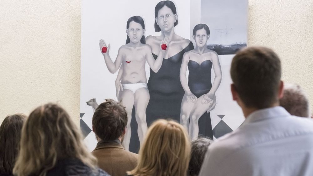 Viewers watch a contemporary painting in which a black-haired woman dressed in black holds two girls on her lap. One of them shows the inside of her hands, which have stigmata on them.