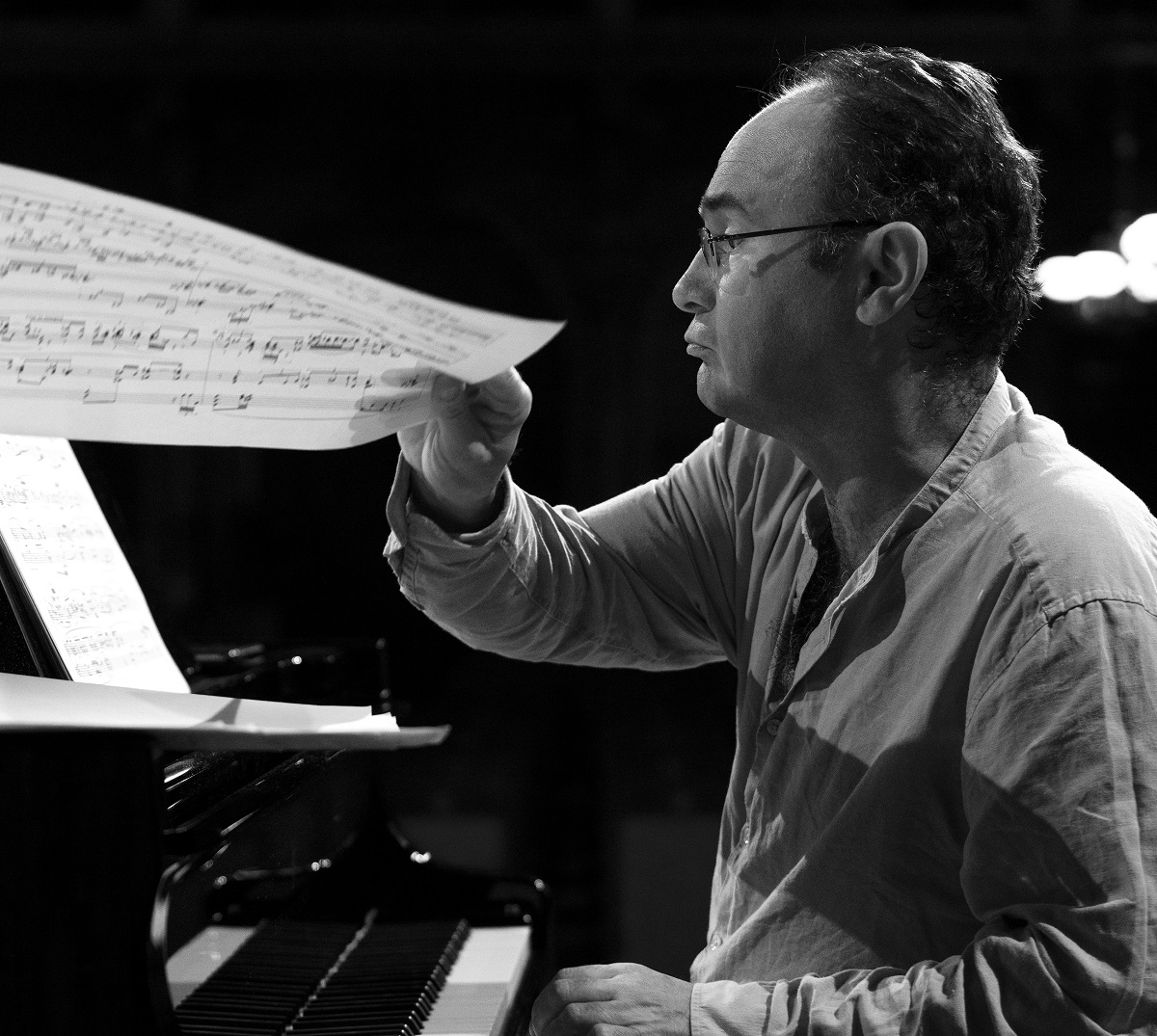 Black and white photo of a man sitting at the piano. He holds paperpage with music notes in his hand. - grafika artykułu