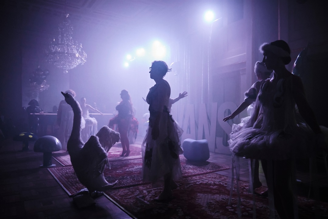 A photo from the show in blue and violet colors. A few women in ballerina costumes on stage. In front of the stage there is a swan with spread wings, above the stage there are spotlights illuminating the stage. - grafika artykułu