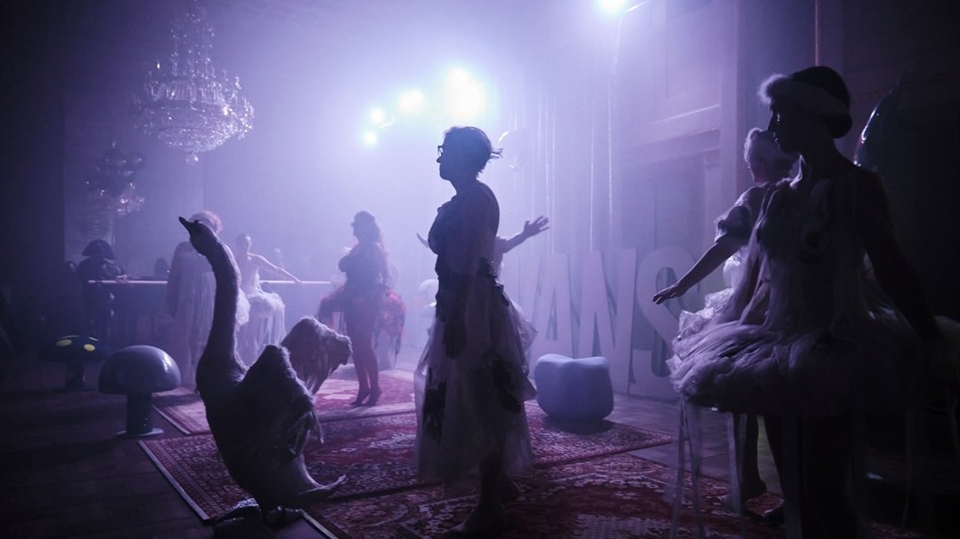 A photo from the show in blue and violet colors. A few women in ballerina costumes on stage. In front of the stage there is a swan with spread wings, above the stage there are spotlights illuminating the stage.