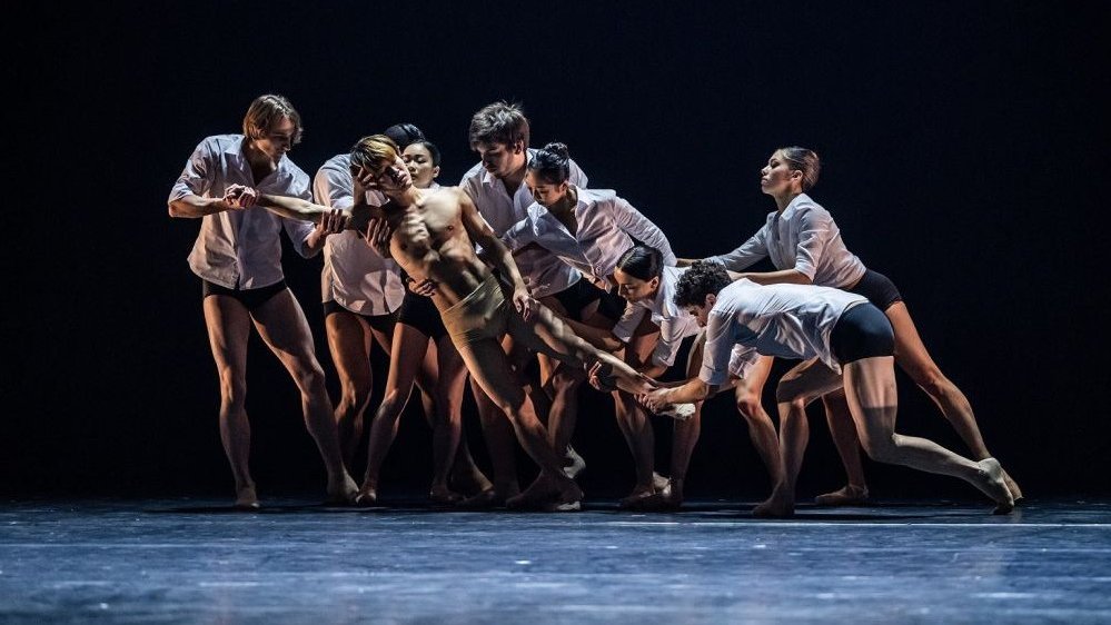 A group of dancers in white shirts hold another dancer, who is performing on stage in his underpants, by his body and head.