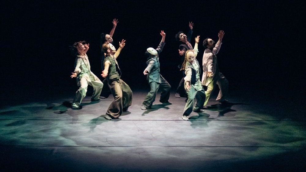 A group of dancers in white face masks dance on stage, raising their hands up. Dark background.