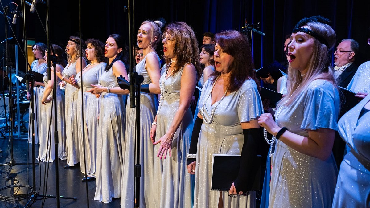 Photo from the concert: women in long white-silver dresses singing on stage.