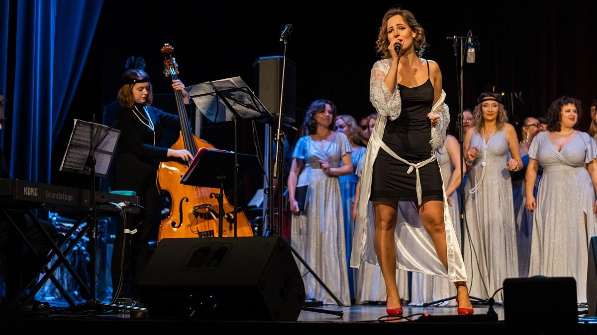 Photo from the concert: a woman singing, on her left a woman playing the double bass, behind her women in long, silver dresses.