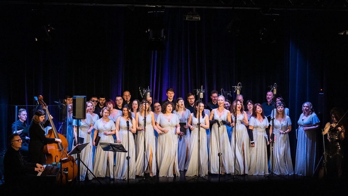 Photo from the concert: women in long, silver and white dresses and several men singing on stage, on the left a woman playing the double bass.