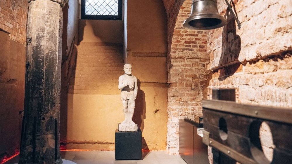A statue of a knight, next to it there was a pedestal on which a sculpture stood. On the right, a medium-sized bell hangs on the brick wall.