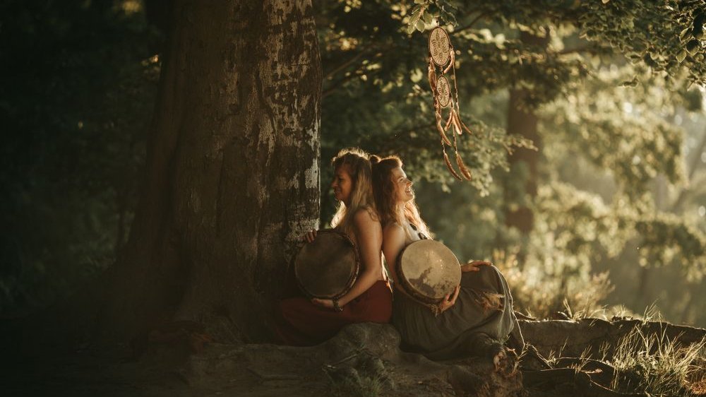 Two girls in dresses are sitting under a large tree, leaning against each other, each holding an old-fashioned drum in her arms. One of them is illuminated by the sun.