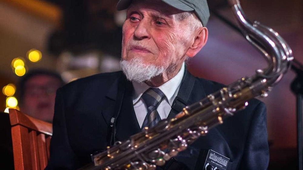 Photo of the elderly man in a peaked cap, holding a saxophone. The man is looking aside.