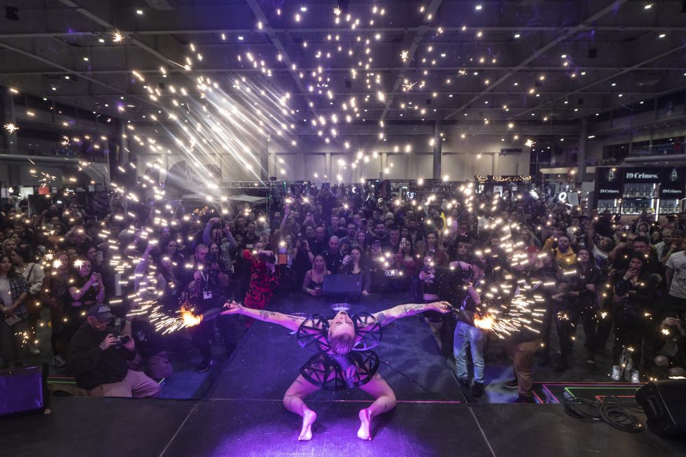 A woman in a futuristic outfit kneels on the stage, with a large audience gathered in front of her. Bright points of light float above people. - grafika artykułu