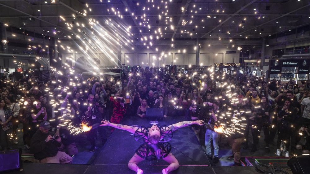 A woman in a futuristic outfit kneels on the stage, with a large audience gathered in front of her. Bright points of light float above people.