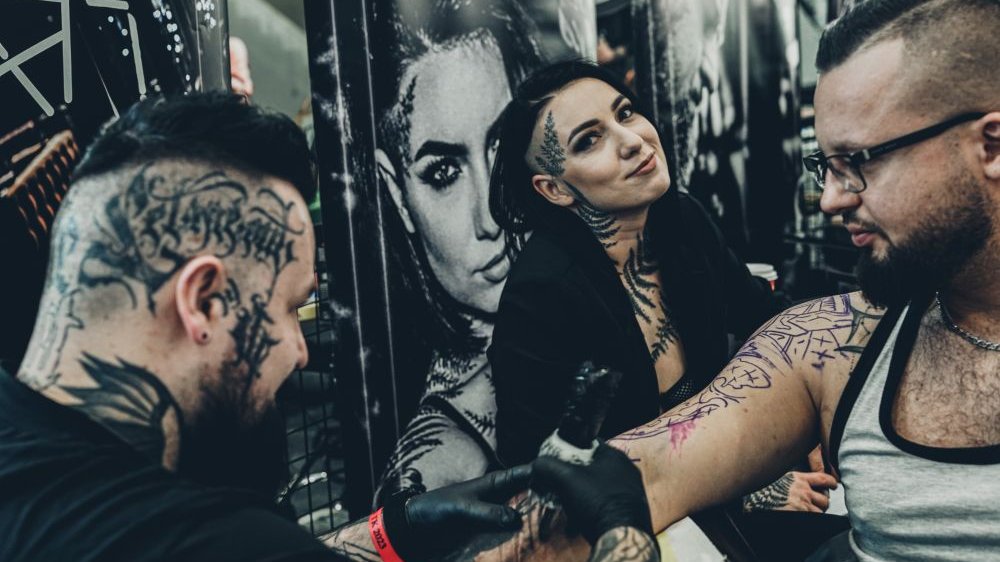 Two men and one woman, one of men works as a tattoo artist. The woman smiles to a camera, the men are busy tattooing.