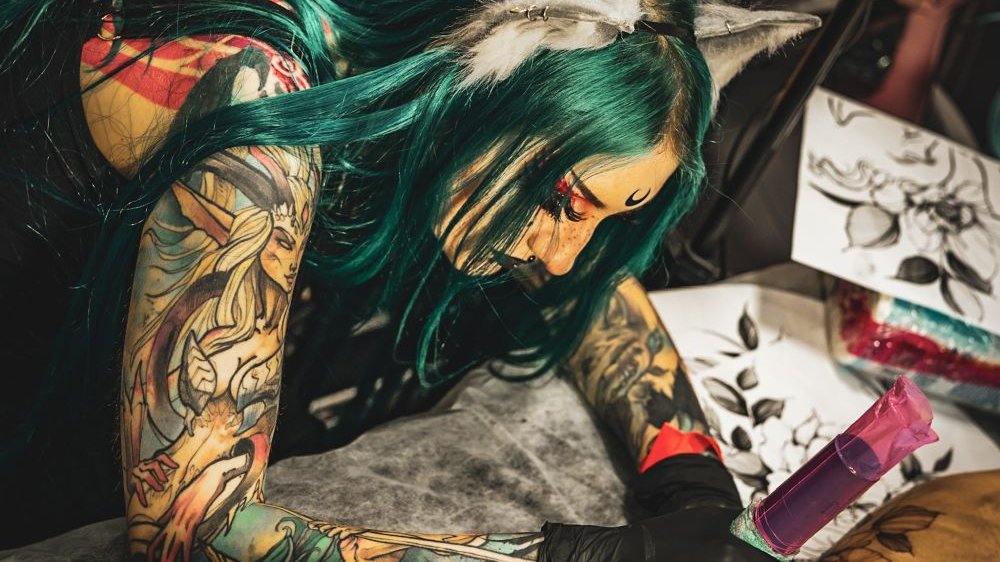 A tattooed girl with green hair and heavy makeup is tattooing a client.