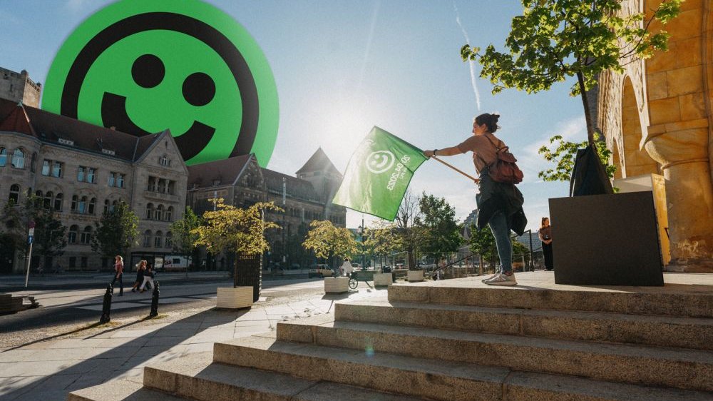 A woman with a green flag in her hands stands on the steps in front of the building. In the background there is a street and a historic building with a smiling face emoticon sticking out from behind it.