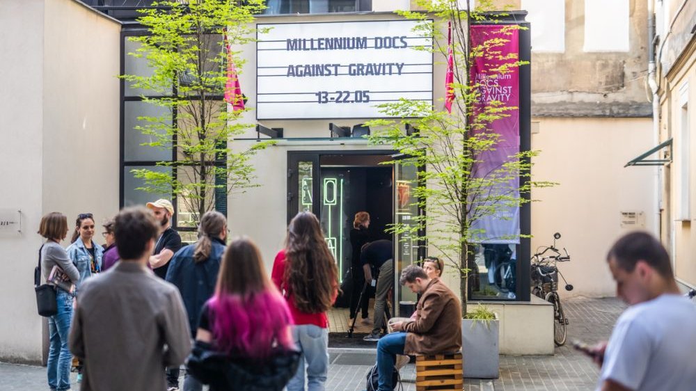 A group of people in front of the entrance to the Muza cinema. Above the door there is a board with the name and dates of the festival.