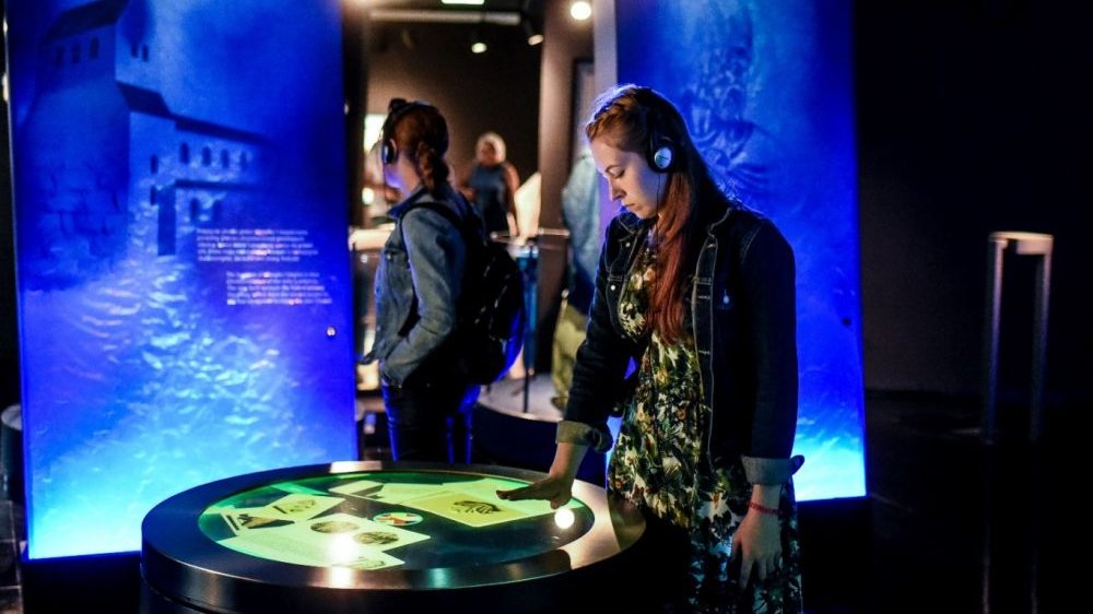 A girl with headphones on looks at the illuminated tabletop with documents displayed on it. Photo in blue colors.
